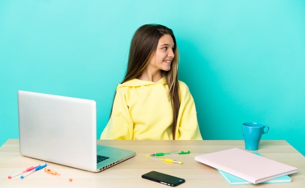 Niña en una mesa con un portátil sobre fondo azul aislado mirando hacia el lado