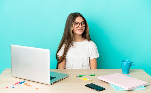 Niña en una mesa con un portátil sobre fondo azul aislado pensando en una idea mientras mira hacia arriba