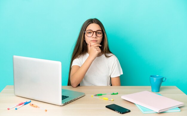 Niña en una mesa con un portátil sobre fondo azul aislado pensando