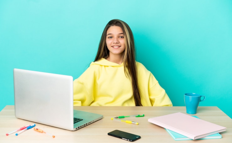 Foto niña en una mesa con un portátil sobre fondo azul aislado posando con los brazos en la cadera y sonriendo