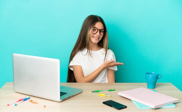 Niña en una mesa con un portátil sobre fondo azul aislado presentando una idea mientras mira sonriendo hacia