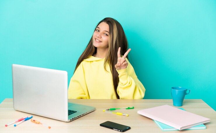 Foto niña en una mesa con un portátil sobre fondo azul aislado sonriendo y mostrando el signo de la victoria