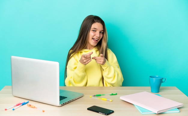 Niña en una mesa con un portátil sobre fondo azul aislado sorprendido y apuntando hacia el frente