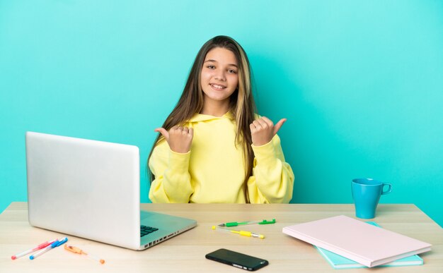 Niña en una mesa con un portátil sobre pared azul aislada con gesto de pulgar hacia arriba y sonriendo