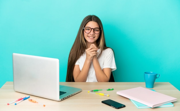 Niña en una mesa con un portátil sobre pared azul aislada riendo