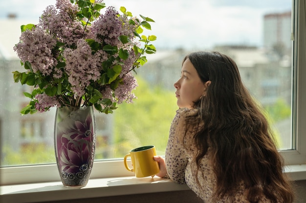 niña mira las flores y bebe té o café de una taza amarilla junto al alféizar de la ventana hay un jarrón con un ramo de lilas