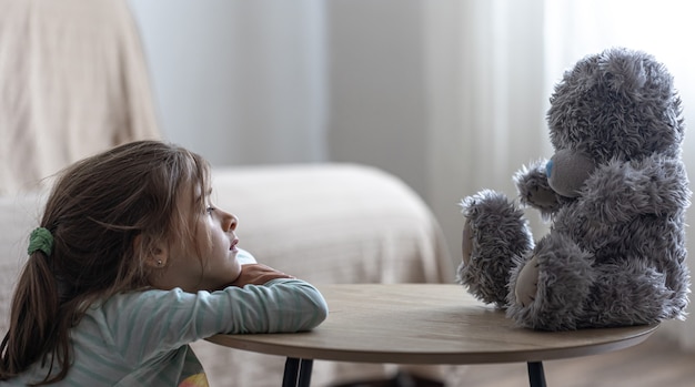 Foto niña mira a su osito de peluche, un niño con un juguete favorito en un espacio de copia de fondo borroso.