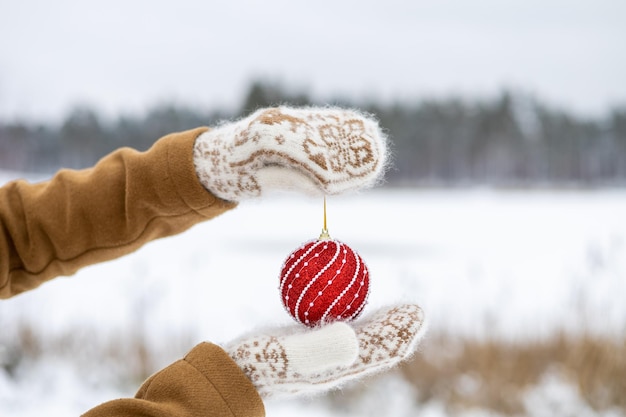 Una niña con mitones de lana sostiene una bola roja de Navidad festiva para el árbol de Año Nuevo en sus manos Desenfoque del bosque