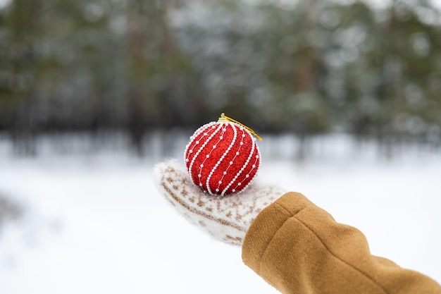 Una niña con mitones de lana sostiene una bola roja de Navidad festiva para el árbol de Año Nuevo en sus manos Desenfoque del bosque