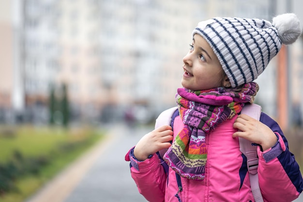 Niña con una mochila en una chaqueta y un sombrero cerca del espacio de copia de la escuela