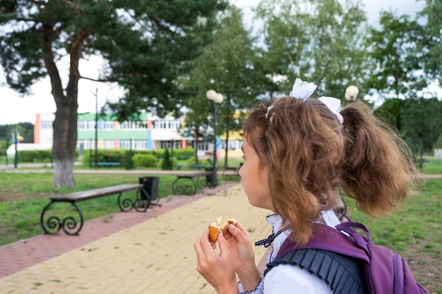 Niña con una mochila comiendo pastel cerca de la escuela Un refrigerio rápido con un panecillo comida poco saludable almuerzo de la escuela Regreso a la escuela Educación clases de escuela primaria 1 de septiembre