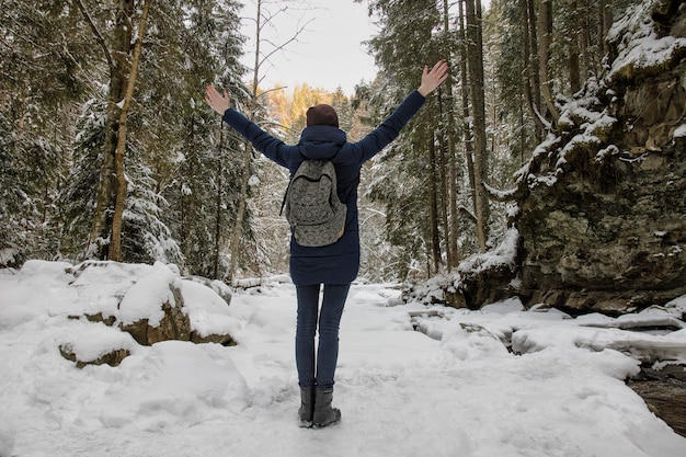 Niña con una mochila está de pie con los brazos levantados en un bosque nevado