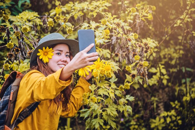 Una niña con una mochila está usando el teléfono para tomar una selfie una foto de la flor amarilla de Bua Tong.