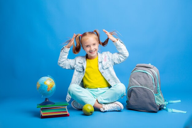 Una niña con una mochila y libros está sentada sobre un fondo azul.
