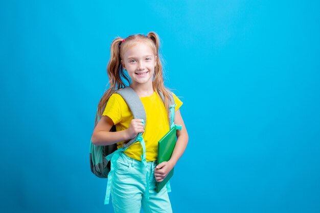 Una niña con una mochila sostiene un libro sobre un fondo azul.