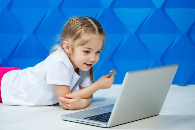 Una niña moderna usa un portátil tirado en el suelo con las piernas mirando a la cámara. Niño gracioso sobre un fondo de pared azul brillante. Tecnología inalámbrica de internet moderna