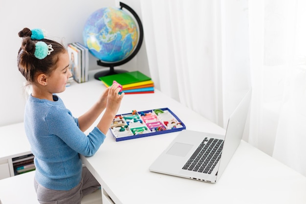 Niña moldeando arco iris de colores de arcilla de juego de niños viendo la lección de aprendizaje en línea en la computadora portátil de interior. Concepto de aprendizaje a distancia en casa.