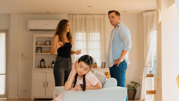 Foto niña molesta e infeliz sentada en un sofá atrapada en medio de la tensión por la discusión de sus padres en la sala de estar. un estilo de vida doméstico poco saludable y una infancia traumática desarrollan depresión. sincronos