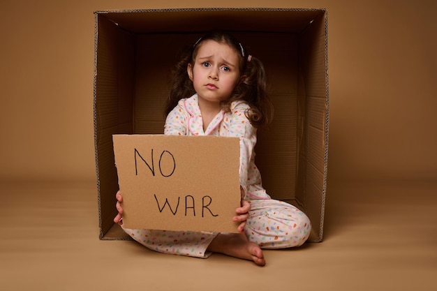 Una niña molesta se sienta dentro de una caja de cartón y muestra un cartel de Stop the War Anuncio social y concepto de alto el fuego los problemas de los migrantes y refugiados durante el conflicto militar