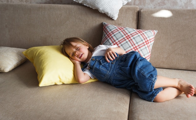 Una niña en un mono de mezclilla azul duerme en casa en el sofá entre almohadas de colores