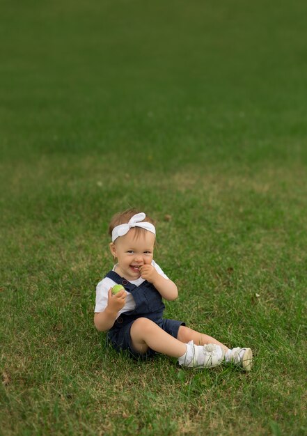 Una niña con un mono de mezclilla se sienta en el parque en el césped y come una manzana