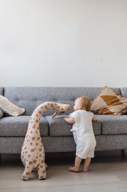 Una niña con un mono de muselina se encuentra con un juguete de jirafa de peluche cerca de un sofá gris en la sala de estar