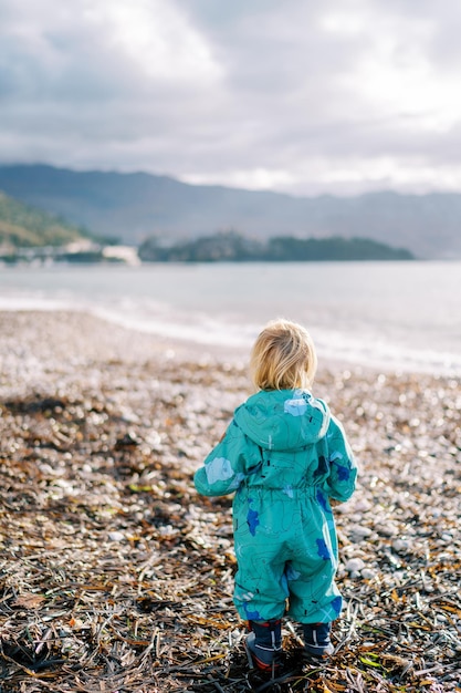 Niña en mono de pie en las algas en la playa y mira el mar hacia atrás