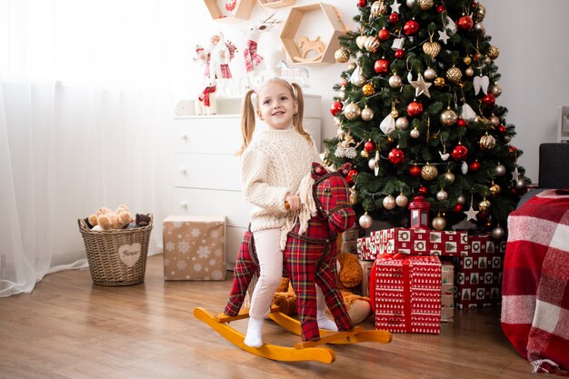 Niña montando a caballo de juguete en casa cerca del árbol de Navidad y cajas de regalo