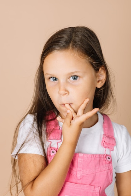 Niña mordiéndose las uñas con ojos brillantes mirando a la cámara con un mono rosa brillante y w...