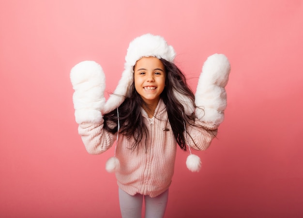 Niña morena con el pelo largo en un gorro de invierno y guantes sobre un fondo rosa