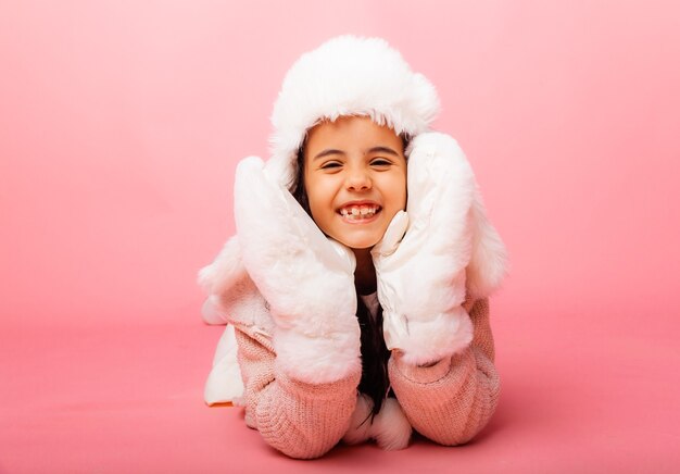 Niña morena con pelo largo en un sombrero de piel de invierno y guantes sobre un fondo rosa.