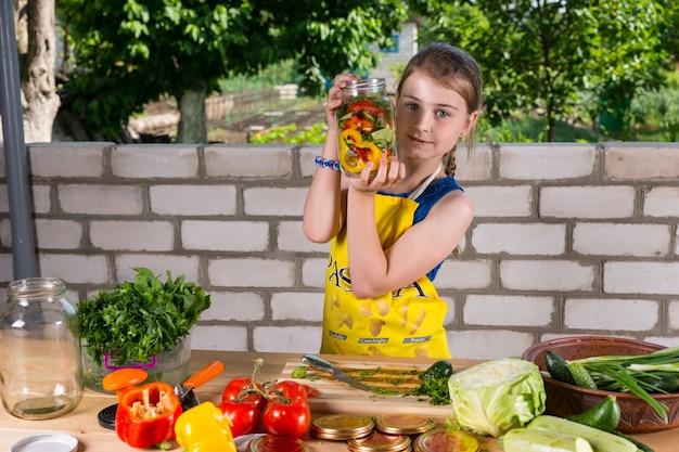 Niña mostrando una botella de verduras frescas que acaba de terminar de cortar en cubitos y colocar en el frasco de un surtido de verduras frente a ella