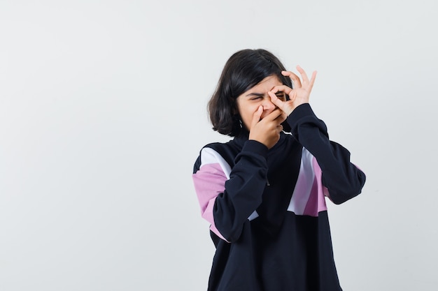 Niña mostrando signo ok en el ojo mientras bosteza en camisa, vista frontal.