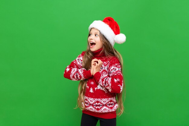 Una niña muestra deleite con un regalo de Navidad Un niño con el pelo largo en un suéter de reno y un sombrero de Santa Claus sobre un fondo verde aislado