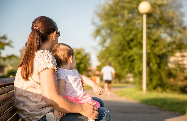 Niña y mujer con coleta sentada en un banco del parque mirando al corredor del hombre