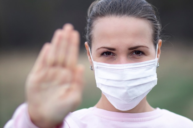 Niña, mujer joven con máscara médica estéril protectora en su rostro al aire libre, muestra la palma, la mano, no deje ningún signo. Contaminación del aire, virus, concepto de coronavirus pandémico. COVID-19