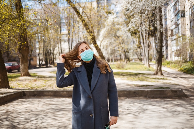 Niña, mujer joven con una máscara médica protectora estéril en su rostro, mirando a la cámara al aire libre, en el jardín de primavera. Contaminación del aire, virus, concepto de coronavirus pandémico.