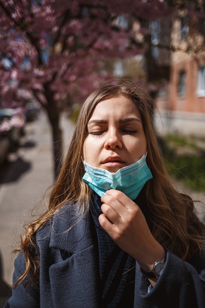 Niña, mujer joven con una máscara médica protectora estéril en su rostro, mirando a la cámara al aire libre, en el jardín de primavera. Contaminación del aire, virus, concepto de coronavirus pandémico.