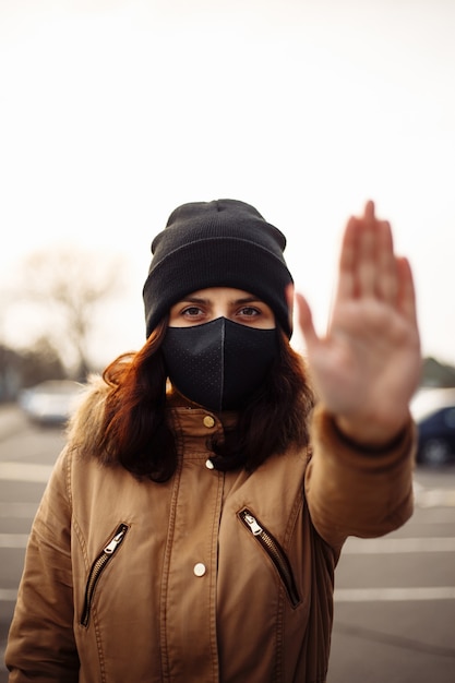 Niña, mujer joven con máscara negra médica estéril protectora en su rostro mirando a la cámara al aire libre, en la calle de la ciudad muestra la palma, la mano, no pare ninguna señal. Contaminación, virus, concepto de coronavirus pandémico.