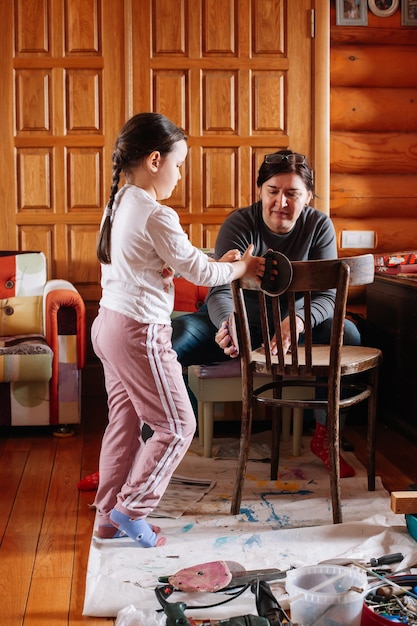 Niña y mujer de mediana edad quitando la vieja capa de pintura de la silla con papel de lija en un taller brillante para la renovación de muebles Prácticas sostenibles Reutilización de cosas viejas Reciclar