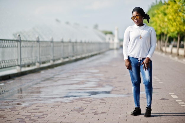 Niña musulmana africana con gafas de sol, hiyab negro, sudadera blanca y jeans, posada al aire libre contra la fuente