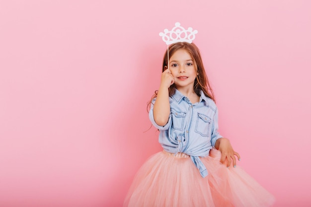 Niña muy dulce con cabello largo morena en falda de tul con corona blanca en la cabeza aislada sobre fondo rosa. Hermoso niño alegre que expresa verdaderas emociones. Lugar para el texto