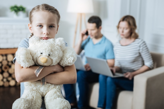 Niña muy infeliz abrazando a su osito de peluche y sintiéndose sola mientras sus padres trabajan en sus computadoras portátiles