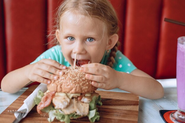 Una niña muy linda con cabello rubio y ojos azules está tratando de morder una hamburguesa grande en un café local. Retrato.