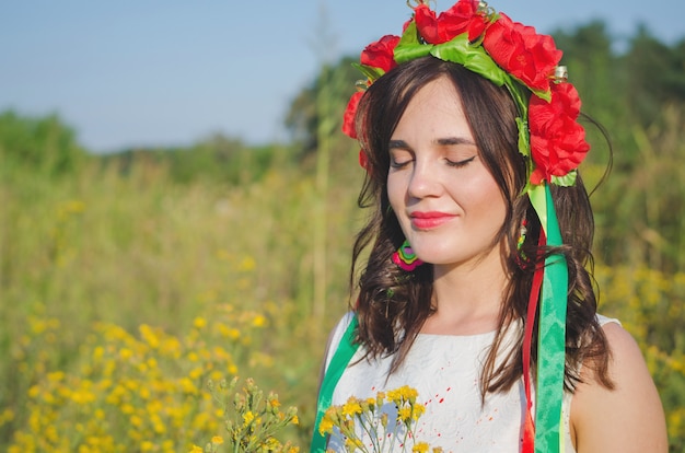 Niña muy sonriente en una guirnalda de flores de amapola y vestido étnico bordado nacional posando en el prado de flores de campo amarillas iluminadas con sol de verano