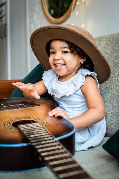 Foto una niña negra con un sombrero se sienta en un sofá y toca la guitarra que está tocando el bebé.