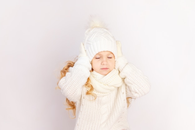 Niña niña con un suéter y sombrero sosteniendo su cabeza con los ojos cerrados en blanco