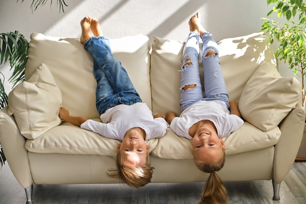 La niña y el niño bonitos están acostados de espaldas en el sofá en la sala de estar, mirando a la cámara y sonriendo mientras juegan en casa