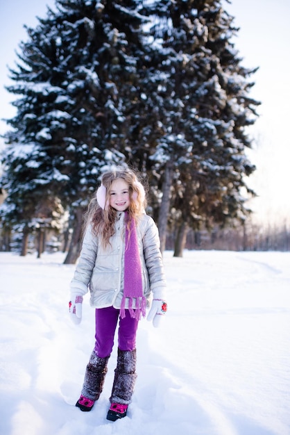 Niña niño divertido lindo 5-6 años a pie en el parque cubierto de nieve sobre fondo de naturaleza al aire libre. El niño pequeño usa chaqueta de invierno, bufanda y auriculares afuera. Infancia.