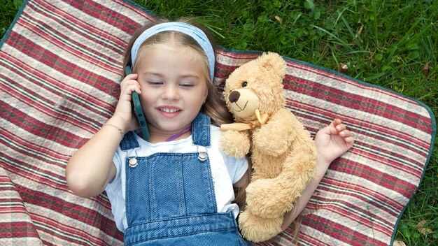 Niña niño feliz acostado sobre una manta sobre césped verde en verano con su osito de peluche hablando por teléfono móvil.